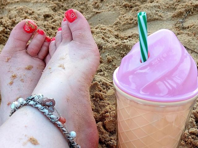 HotSandy4You-sex's sandy feet with red nail polish and heart designs, wearing a chain bracelet, next to a pink ice cream with a green straw on a sandy beach.