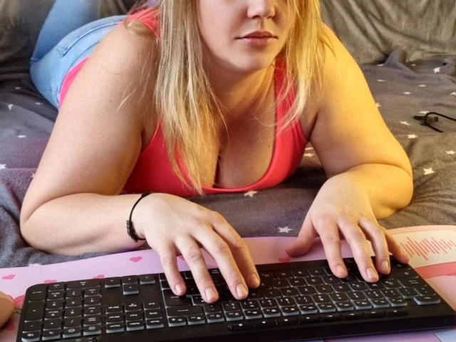 LiesaBDSM-hot with blonde hair, pink tank top, and blue jeans, lying on bed, typing on black keyboard with hands on keys, looking slightly down.