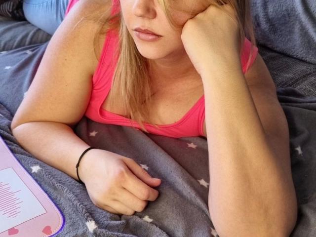 LiesaBDSM-hot with blonde hair, pink tank top, resting chin on hand, lying on grey star-patterned blanket, right wrist with black bracelet, looking down, pink lips, soft expression.