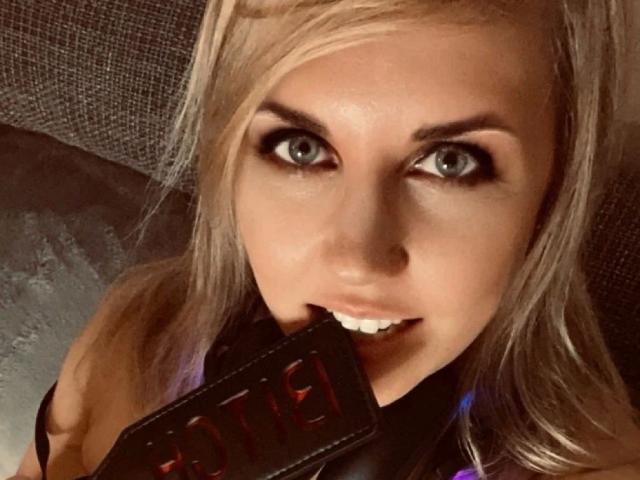 LiesaBDSM-hot with blonde hair, blue eyes, and a sexy smile, biting a black leather strap with "JADE" in red. Her eyes are wide and her lips are parted.