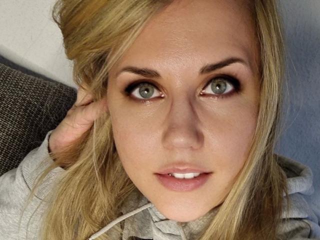 LiesaBDSM-hot has green eyes, blonde hair, and is wearing a grey hoodie, looking directly at the camera with a slightly open mouth and soft pink lips.