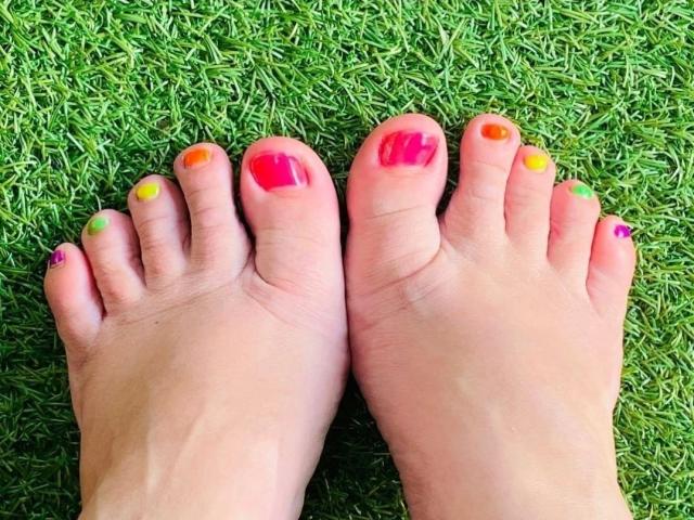 LiesaBDSM-hot's feet with colorful toenails: pink, red, yellow, orange, purple, and pink, standing on green grass.