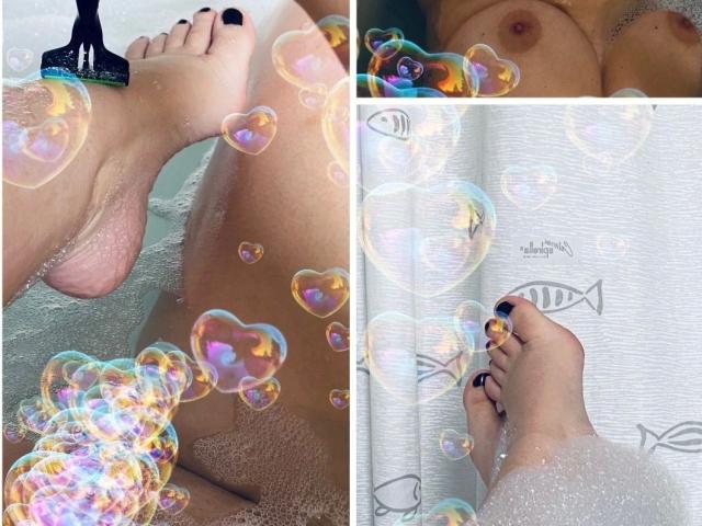 LiesaBDSM-hot's bare feet in a bubble bath, with colorful soap bubbles, black-painted toenails, and her tits visible in the top right. Her toes are against a white curtain.