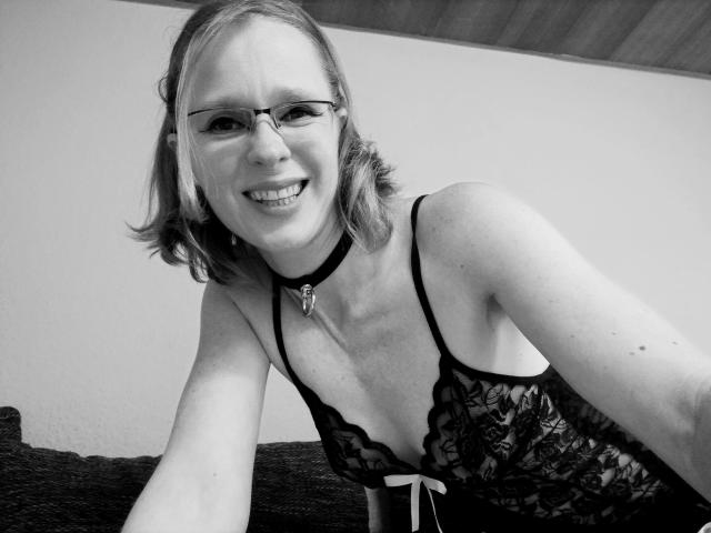 HeisseLina-hot, wearing black lace lingerie with a key choker, smiling at the camera, has small tits, light skin, glasses, and shoulder-length blonde hair, in a black-and-white photo.