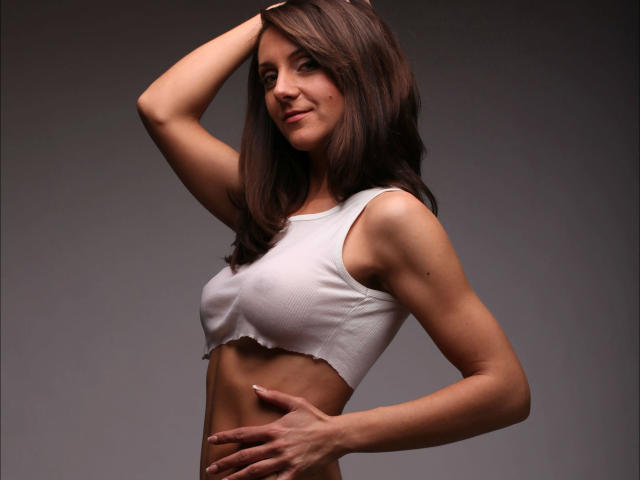 LarisaRais-hot with brown hair, white crop top, small tits, and hand on head, standing against gray background, showing slim, toned, and attractive upper body.