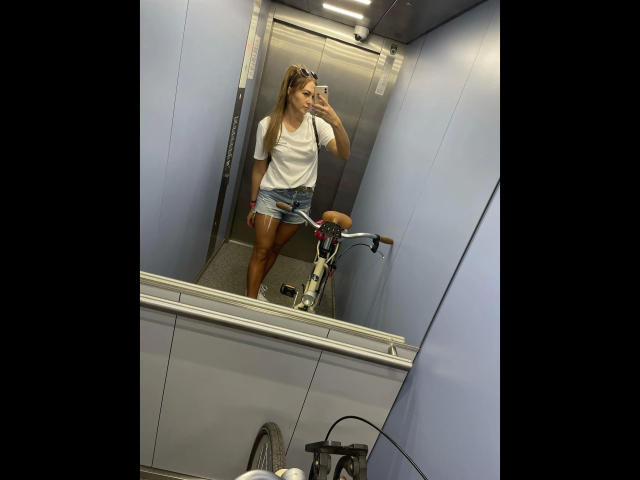 GanzNettePaola-hot steht in einem Aufzug, trägt ein weißes Tanktop, Jeansshorts und hält ein Fahrrad. Sie macht ein Selfie und zeigt ihre gebräunten Beine und kleine, pralle Brüste. Spiegelung im Spiegel..