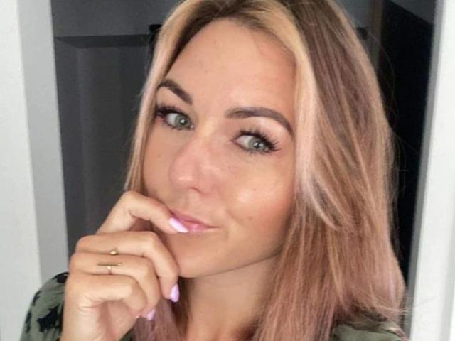 GanzNettePaola-hot with blonde hair, green eyes, and pink lipstick, slightly smiling, touching her chin with a finger, wearing a green top. Her nails are pink, and she has a gold ring.