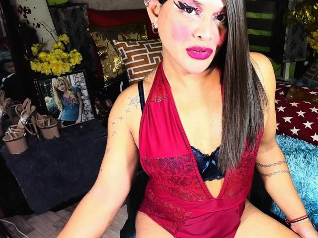 TransGiftedCum with pink makeup, red lace halter, black bra, small tits, tattoos, long brown hair, pink lipstick, sitting, showing pussy, black bra, lace, and blue fluffy pillow.