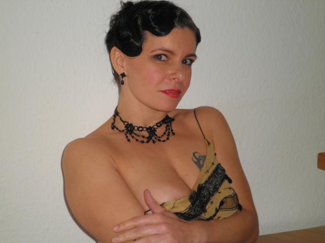 MaitresseGina-hot with black curly hair, red lipstick, black bead necklace, and yellow-black dress covering her tits, arms crossed, tattoo on left side, white wall in background.