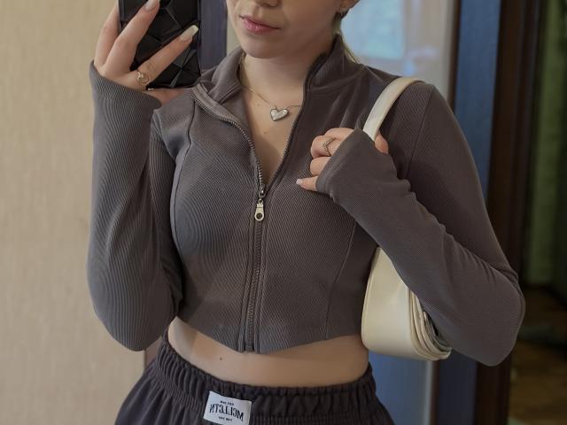 SugarrX, avec une poitrine de taille moyenne, portant un haut gris à fermeture éclair et une jupe noire, tenant un téléphone, un sac blanc sur l'épaule, un collier avec un cœur, fesses légèrement visibles..