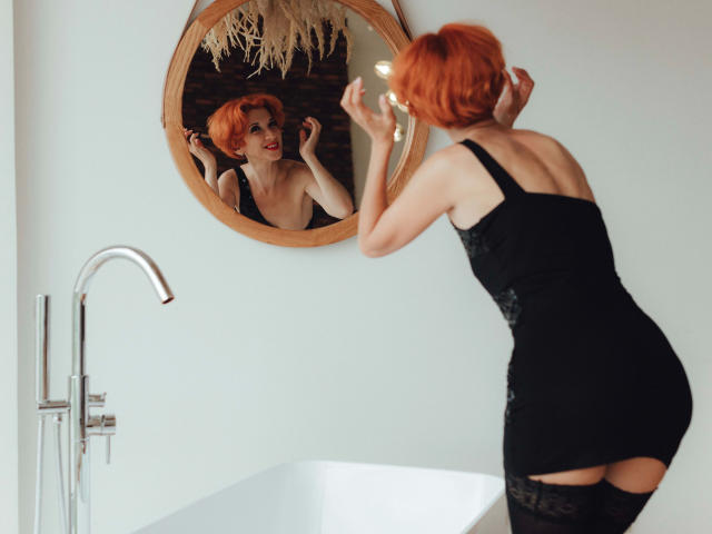 Maike75-hot with short red hair, black lace dress, and thigh-high stockings, looking at herself in a mirror, showing her ass, in a bathroom with a white tub.