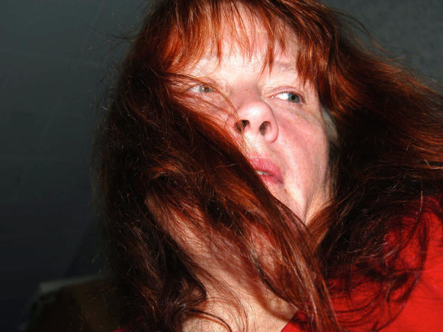 Johannalive, l'air bouillant, avec des cheveux roux en bataille qui lui couvrent le visage, une peau pâle et des yeux bleus, portant une chemise rouge, regardant légèrement sur le côté avec une expression neutre..
