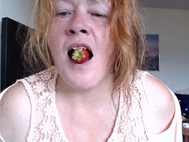 Johannalive-hot has red, messy hair, pale skin, and freckles. She's wearing a lace top, with a strawberry in her open mouth, looking slightly bored or tired.