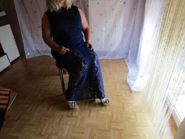 Hotferkel-sex sitting on a chair wearing a blue lace dress, showing her ass, in a room with wooden parquet flooring and white curtains. She's wearing silver high heels.
