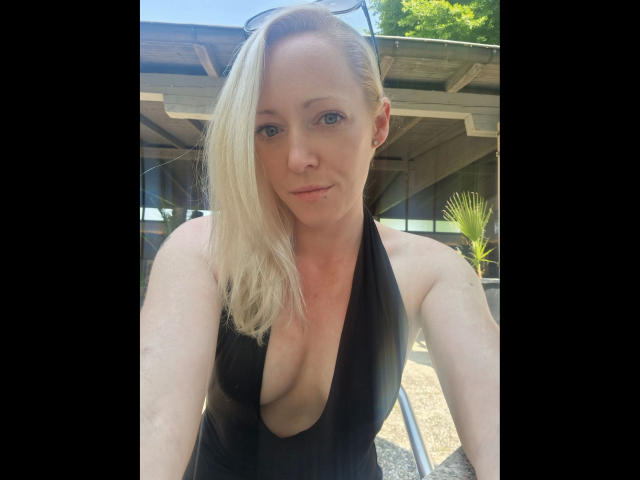 LeaKirsch-hot has blonde hair, blue eyes, and a black one-piece swimsuit with a deep cleavage, showing her large tits. She's smiling, with sunglasses on her head.