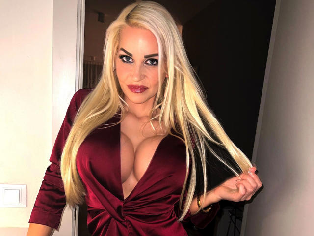 Jackylafey-hot with long blonde hair, deep red dress, and large boobs, holding her hair, staring with blue eyes and red lipstick.