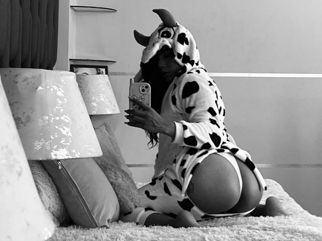Amberv in a cow onesie with horns, kneeling on a bed, showing her ass in white panties, taking a selfie with a phone. Black and white photo.