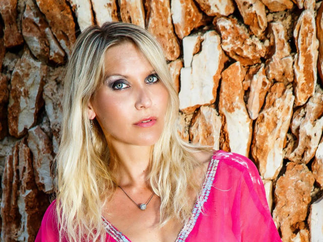 JuliaPink-hot a les cheveux blonds, les yeux bleus, et porte un haut rose transparent avec des détails violets. Elle porte un collier avec une petite pierre précieuse et se tient devant un mur rocheux..