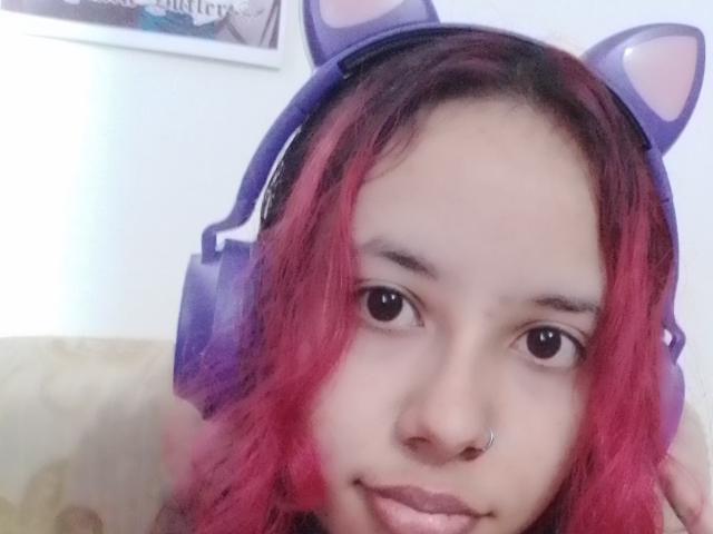 KiaraManson with purple cat-ear headphones, red and pink ombre hair, nose ring, and cute smile, looking directly at the camera. Her ass is not visible.