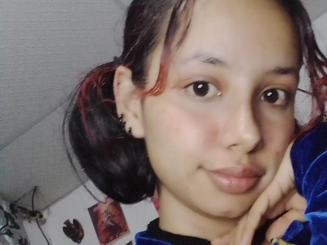 KiaraManson has medium brown skin, black hair with red highlights, and small earrings. She's smiling with her chin resting on her hand, wearing a blue velvet shirt.