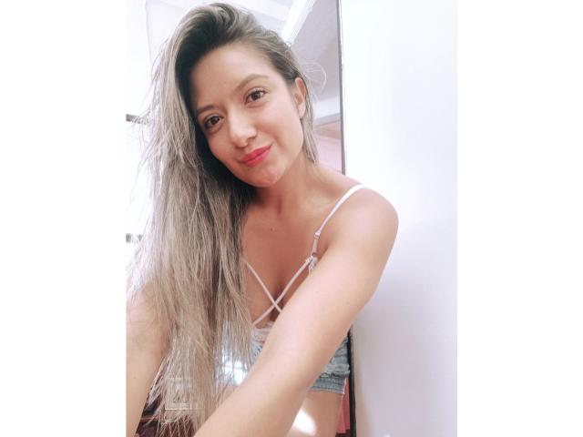 ValeriaHunter69 with long, wet, blonde hair, smiling, wearing a white strappy top, showing small tits, in a white-walled room. She's looking at the camera with red lips.