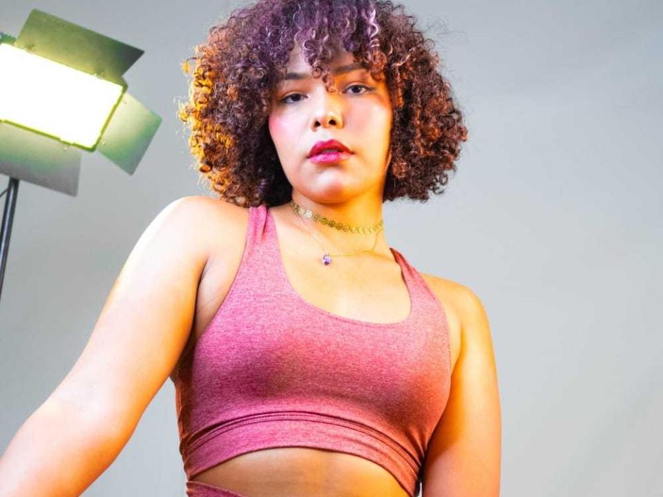 NiaColussi met krullend bruin haar, die een roze sportbeha draagt, kijkt naar beneden met een serieuze uitdrukking, met een zichtbaar roze tepel, en staat in de buurt van een groene lichtbron..