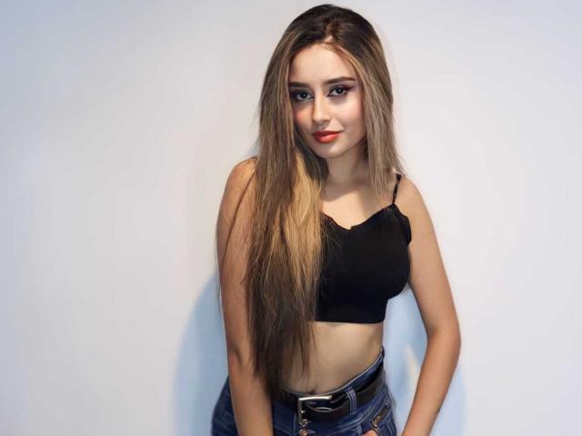 MakennsyWhite with long blonde hair, black crop top, and blue jeans, standing, hands on hips, showing small tits, cute face, red lipstick, and a slight smile.