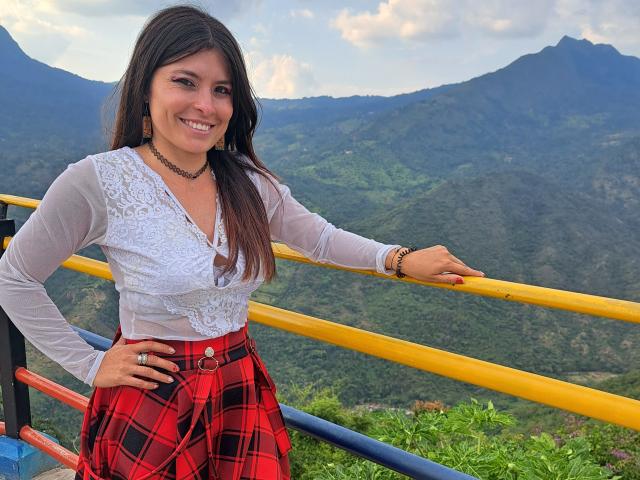 AlexandraSaenz69, avec de longs cheveux bruns, souriante, portant un haut blanc en dentelle, une jupe à carreaux rouges, un collier noir et un bracelet, appuyée sur une rambarde jaune, avec des montagnes vertes en arrière-plan..