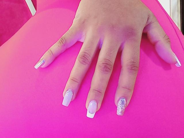La main de JackelineRoset, avec de longs ongles roses, blancs à la pointe et argentés pailletés, repose sur une surface rose. Ses doigts sont écartés, mettant en valeur sa manucure élégante et séduisante..