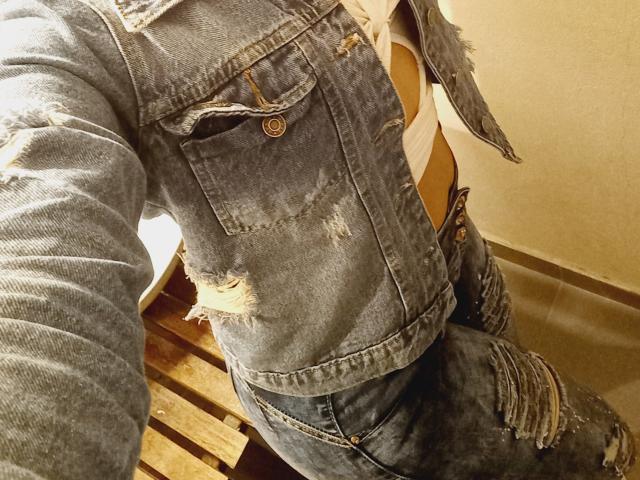 Jackeline Roset vestindo uma jaqueta jeans azul rasgada e calças jeans combinando com bordas desfiadas, mostrando tecido rasgado no peito e na coxa, em pé sobre uma superfície de madeira..