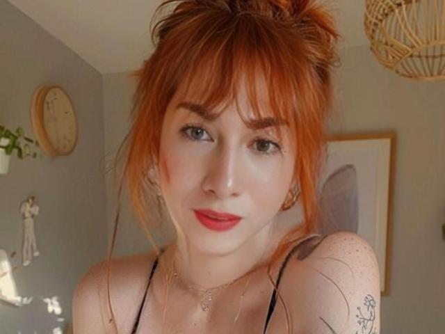 SexyTopHotMistress with red hair, bangs, and freckles, wearing a black top, smiling, red lipstick, small tattoo on right shoulder, and a delicate necklace, in a sunlit room.