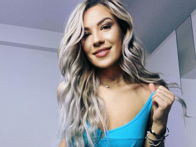 ZoeyMarie with long, wavy blonde hair, pulling down a blue tank top to show her small, perky tits, smiling at the camera. She's wearing a necklace and bracelets.