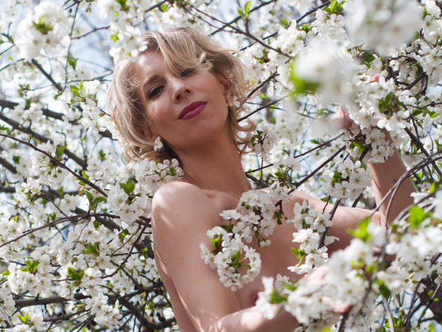 AdrianaSea with curly blonde hair, pink lipstick, and bare shoulders, surrounded by white cherry blossoms, holding branches, looking up with a slight smile. Her tits are partially obscured.