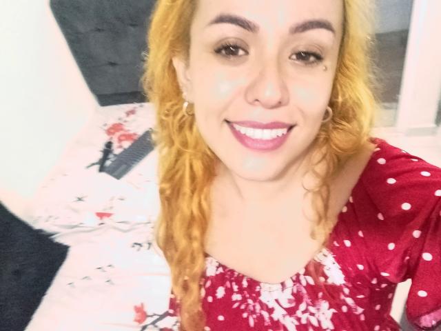 Joanallen with blonde curly hair, smiling, wearing a red polka dot dress, white splatter on face, pink lipstick, hoop earrings, sitting on bed with floral sheets.