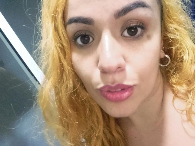 Joanallen a les cheveux bouclés et blonds, les yeux marron, des sourcils épais, du rouge à lèvres rose et une boucle d'oreille argentée. Elle regarde l'appareil photo avec un léger froncement de sourcils..