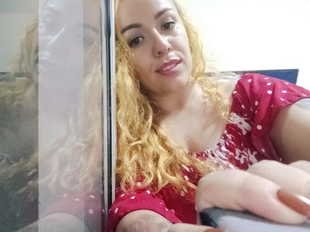 Joanallen, avec ses cheveux blonds bouclés, sa robe rose à pois et son anneau de nez, sourit à l'appareil photo, son bras reposant sur une vitre. Ses seins sont légèrement visibles..