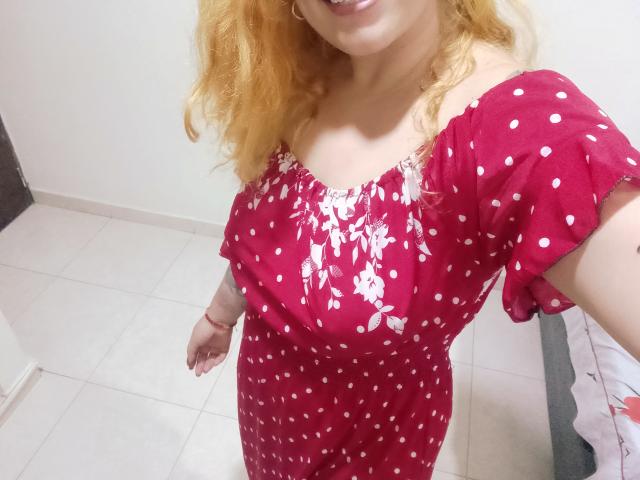 Joanallen, avec ses cheveux blonds bouclés, portant une robe rouge à pois avec des motifs floraux blancs, montrant ses seins nus et souriant, debout sur un sol carrelé blanc..