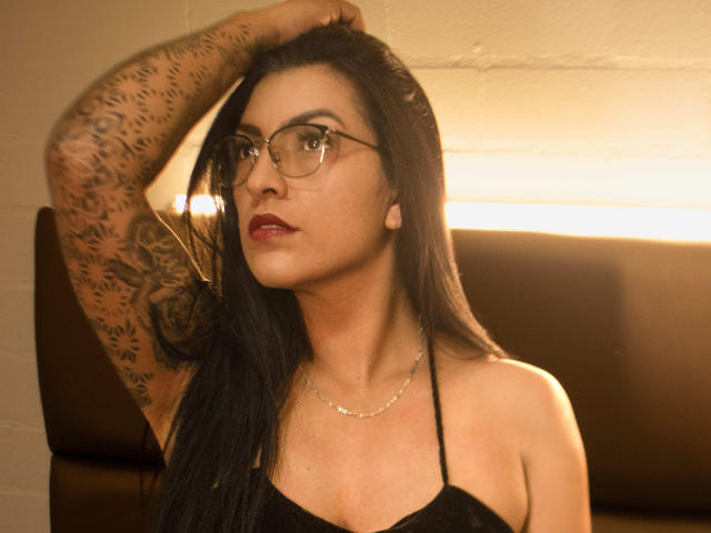 JanetteRider with black hair, glasses, and tattoos on her arm, wearing a black halter top, raises her arm, showing off her tits and a silver necklace.