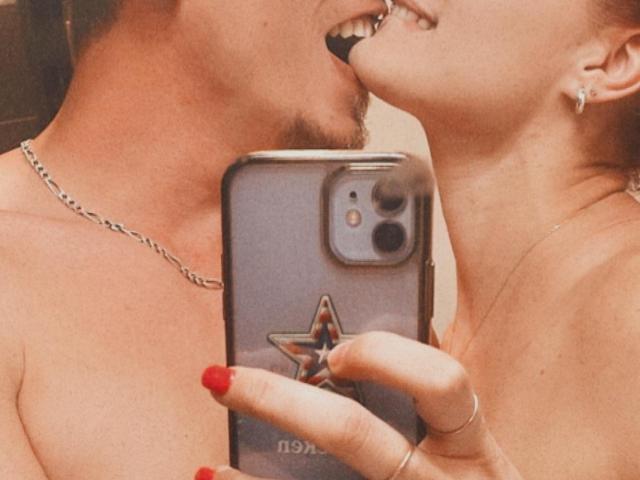 LesLioups met kort bruin haar en een baard, die LesLioups kust met rode nagels en een telefoonhoesje met sterren, waarbij de blote schouders en nekken zichtbaar zijn, en die oorbellen en een ketting draagt..