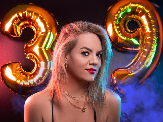 AvaMia with blonde hair, red lipstick, gold necklace, and black top, standing in front of shiny gold "21" balloons. Her tits are small, and she's looking at the camera.