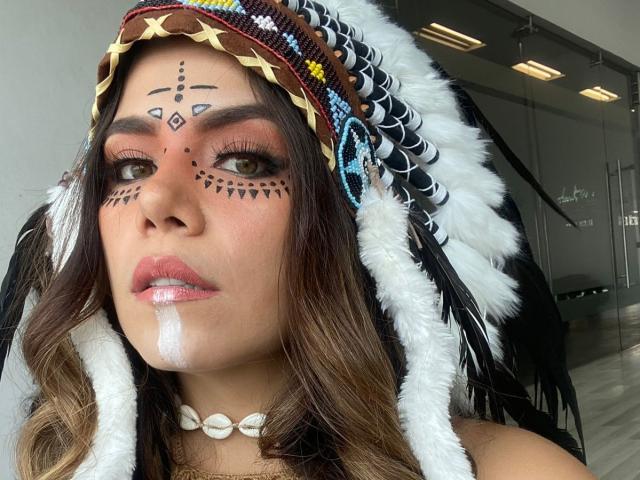 NathalieSwan with brown eyes, face paint, and white facial streak, wearing a feathered headdress, shell necklace, and pink lipstick, looking at the camera. Boobs not visible.