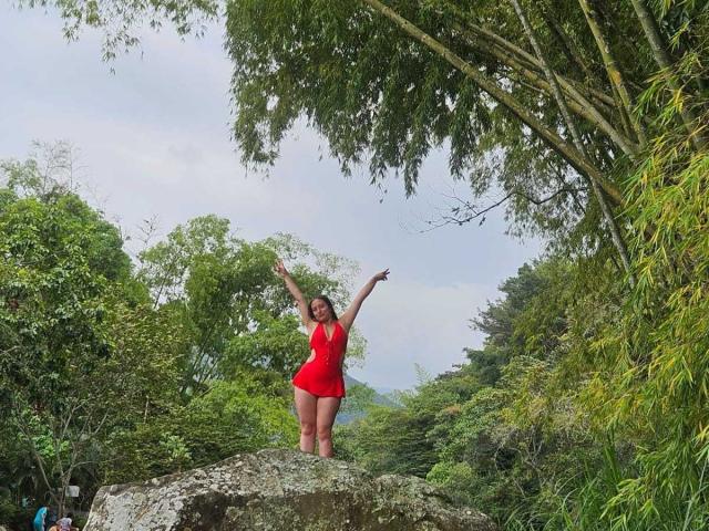 VenusSweetPalmer se tient sur un rocher, les bras levés, vêtue d'une robe rouge, montrant ses fesses, entourée d'arbres verts luxuriants, souriante, avec un ciel nuageux au-dessus..