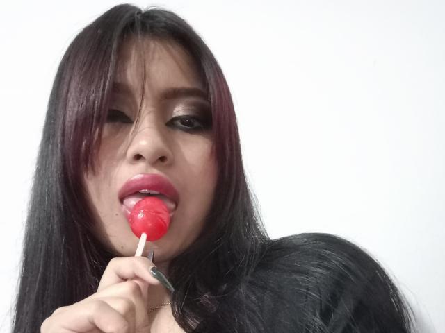 EmmaaRodriguez with long black hair, red lipstick, and dark eye makeup, sucking on a red candy lollipop, holding it with her right hand, showing her glossy pink tongue.