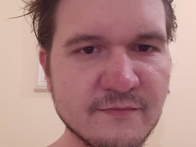 O rosto de NectarineFad, levemente suado, com uma pequena barba e bigode, cabelo castanho ondulado e um sorriso discreto, mostrando um leve rubor no nariz e nas bochechas..