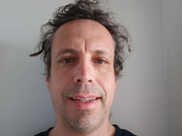 JacostaX tiene una frente ligeramente arrugada, cabello oscuro y rizado, piel clara, ojos marrones y una sonrisa sutil con dientes visibles. Lleva una camisa oscura..