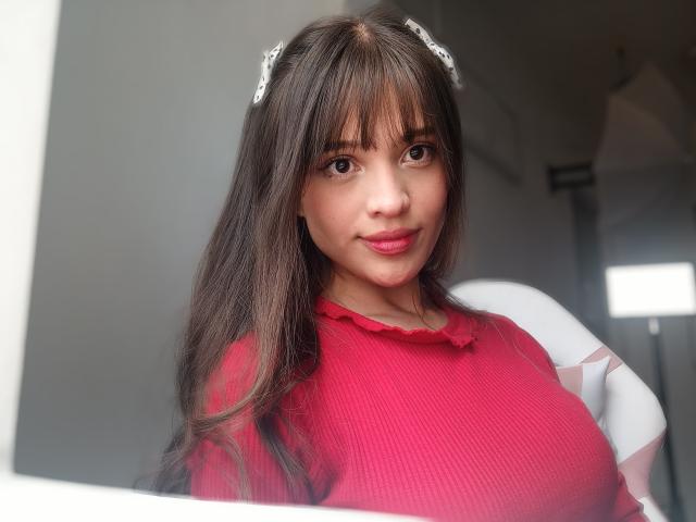 MelodySwet with long brown hair, bangs, and white hair clips, wearing a red ribbed top, showing her big tits, smiling, standing by a window, sunlight on her face.