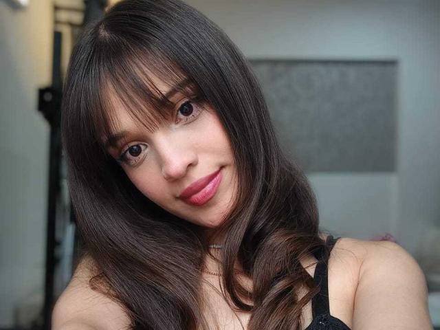 MelodySwet has long brown hair, bangs, and pink lips. She's smiling with brown eyes, wearing a black strap, and has a cute, sexy face.