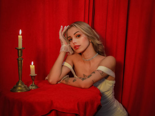 Lara Moss, avec ses cheveux blonds, son rouge à lèvres rouge, sa robe blanche à épaules dénudées, des tatouages sur le bras, une main sur le visage, deux bougies allumées sur un tissu rouge, un rideau rouge en arrière-plan..