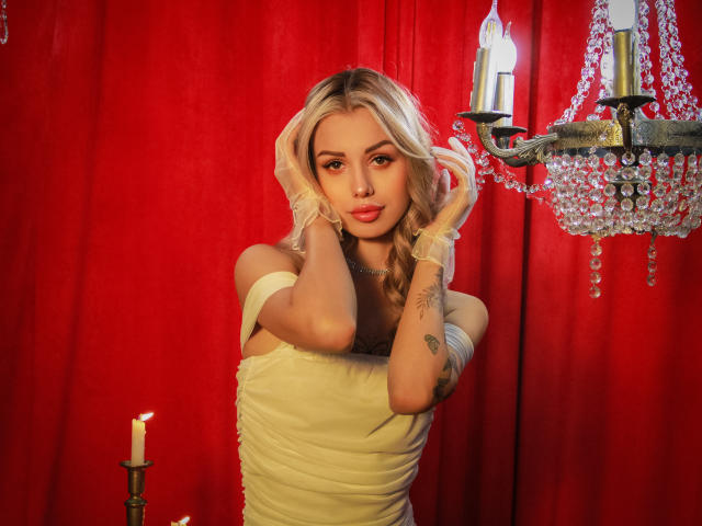 Lara Moss, con cabello rubio, vestida con un vestido amarillo de un solo hombro, guantes blancos y un collar, tocándose las orejas, de pie frente a una cortina roja con un candelabro de cristal..