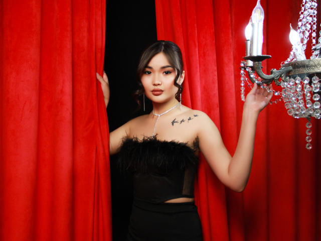 TeyaFaber with dark hair, black feathered top, and high-waisted black pants, holding red curtains, wearing a diamond necklace, and holding a crystal chandelier. Her tits are visible.